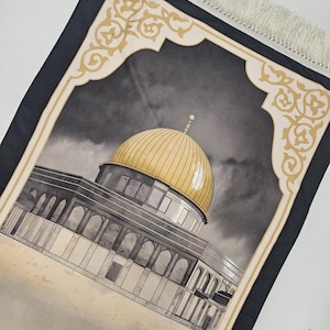 May include: A prayer mat with a gold-domed building illustration, framed by a cream and gold border. The rug has a dark grey background and a cream-coloured fringe. The word "TAWAKKUL" is printed at the bottom.