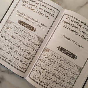 May include: An open book with brown text on white pages. The text on the left page reads "By reading 2 times it is equivalent to the sawaab of reading 1 Qur'an." The text on the right page reads "By reading 2 times it is equivalent to the sawaab of reading 1 Qur'an."