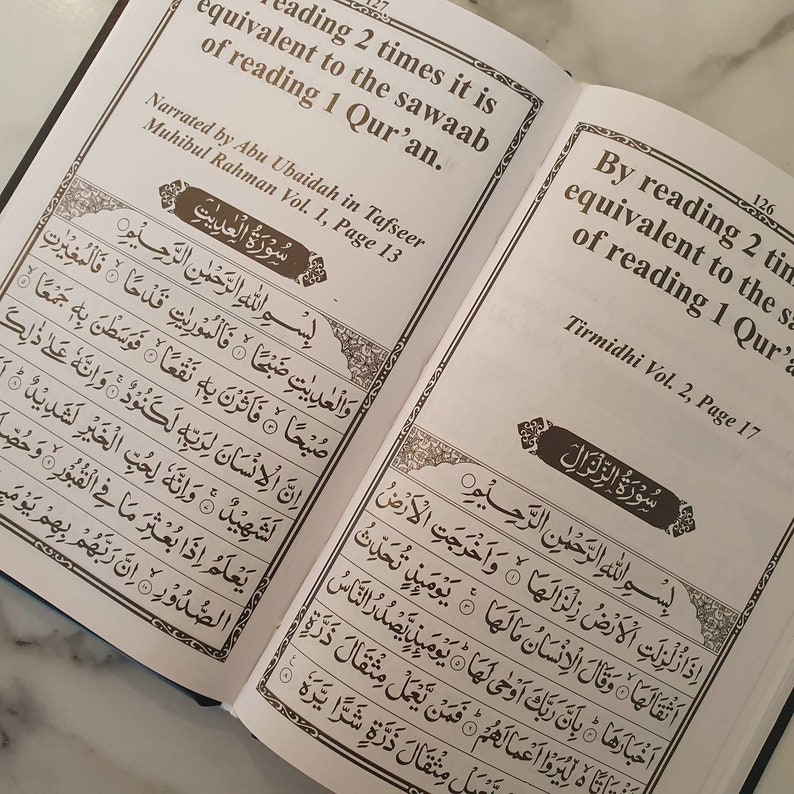 May include: An open book with brown text on white pages. The text on the left page reads "By reading 2 times it is equivalent to the sawaab of reading 1 Qur'an." The text on the right page reads "By reading 2 times it is equivalent to the sawaab of reading 1 Qur'an."