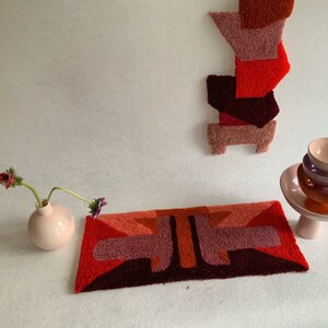 Pu&ograve; includere: Un tappeto geometrico con uno schema di colori rosso, arancione e viola. Il tappeto &egrave; realizzato in un materiale morbido e soffice e ha un design moderno. Un vaso con fiori si trova sul lato sinistro del tappeto, e un set di ciotole si trova su un supporto sul lato destro del tappeto.