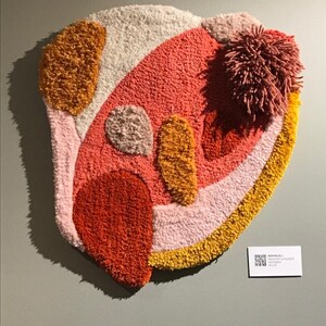 May include: A colorful abstract wall hanging made of fluffy yarn in shades of pink, orange, and yellow. The piece has a unique, organic shape and a textured surface.