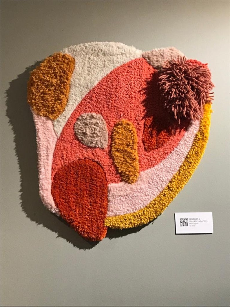 May include: A colorful abstract wall hanging made of fluffy yarn in shades of pink, orange, and yellow. The piece has a unique, organic shape and a textured surface.