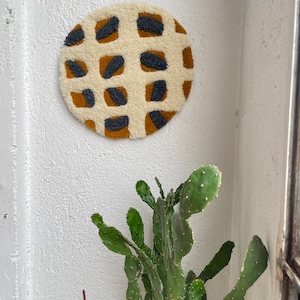 May include: A round wall hanging with a geometric pattern in shades of white, brown, and gray. The pattern is made up of squares and circles.