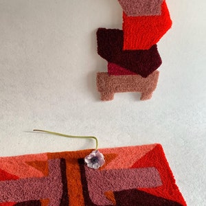 Pu&ograve; includere: Un tappeto geometrico con uno schema di colori rosso, arancione e rosa. Il tappeto &egrave; realizzato in un materiale soffice e testurizzato. Un piccolo fiore rosa &egrave; posizionato sul tappeto.