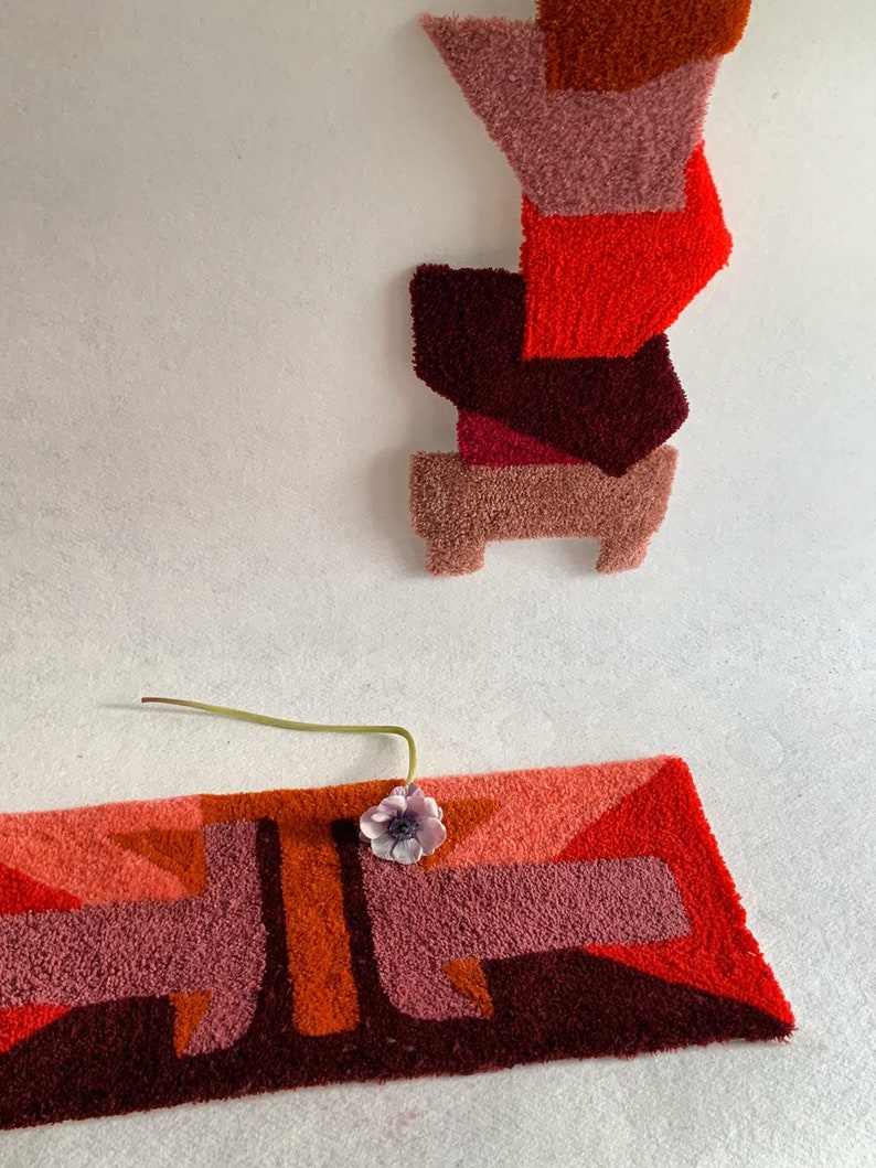 Pu&ograve; includere: Un tappeto geometrico con uno schema di colori rosso, arancione e rosa. Il tappeto &egrave; realizzato in un materiale soffice e testurizzato. Un piccolo fiore rosa &egrave; posizionato sul tappeto.