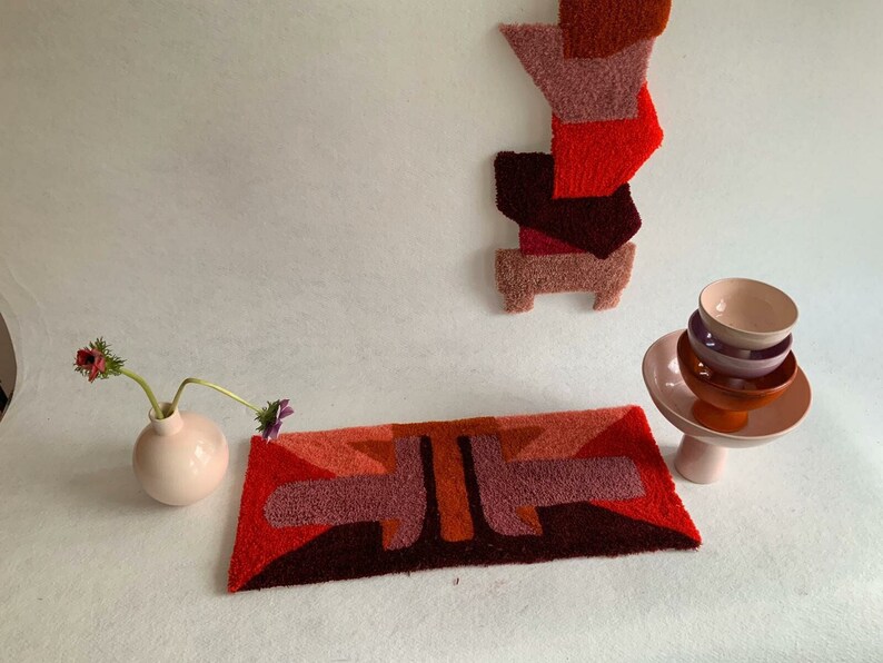 Pu&ograve; includere: Un tappeto geometrico con uno schema di colori rosso, arancione e viola. Il tappeto &egrave; realizzato in un materiale morbido e soffice e ha un design moderno. Un vaso con fiori si trova sul lato sinistro del tappeto, e un set di ciotole si trova su un supporto sul lato destro del tappeto.