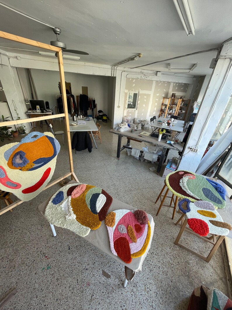 May include: Three colorful, abstract, hand-tufted rugs with different geometric shapes and patterns. The rugs are made with a variety of colors, including red, orange, yellow, green, blue, and pink. The rugs are displayed on a table and a stool in a workshop setting.