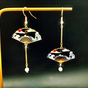 Może przedstawiać: Złote kolczyki wiszące z motywem wachlarza. Posiadają czerwoną słońce, chmury i fale, z niebieskimi, białymi i złotymi detalami. Każdy kolczyk ma wiszącą perłę i jest zawieszony na złotym haczyku.