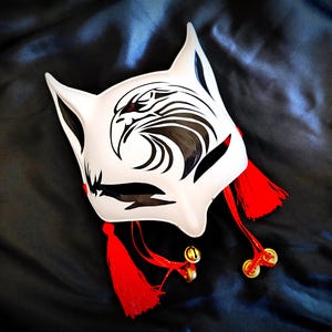 以下が含まれることがあります： 白と黒と赤のアクセントが施された狐のマスク。マスクは、様式化された鳥のデザインと、小さな金色のベルが付いた赤いタッセルが特徴です。マスクは、暗く質感のある背景に配置されています。