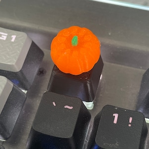 May include: Orange pumpkin-shaped keycap with a green stem, sitting on a black keyboard with the keys "TAB" and "1!" visible.