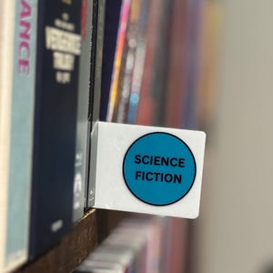 May include: A white rectangular shelf divider featuring a circular blue sticker with the black text SCIENCE FICTION printed in the center. The divider is positioned between two rows of books on a wooden shelf, serving as a genre label.