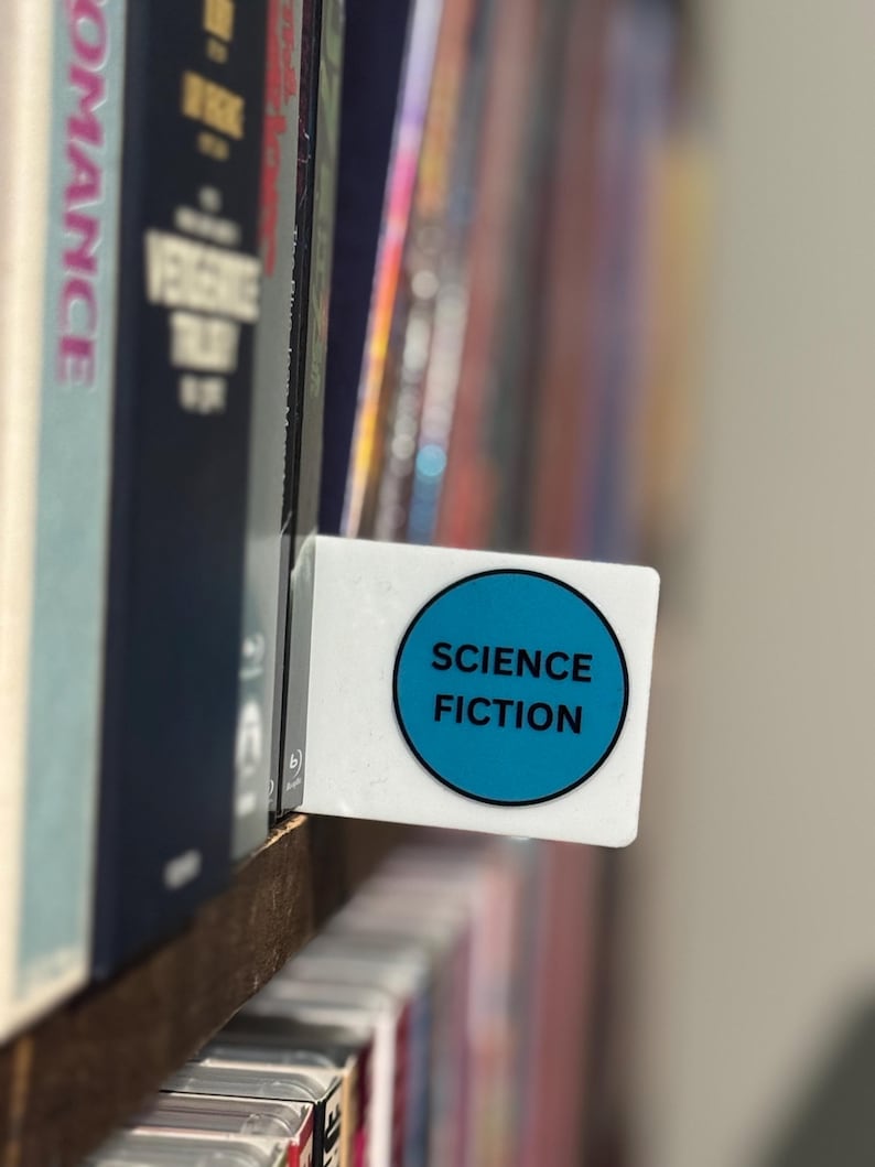 May include: A white rectangular shelf divider featuring a circular blue sticker with the black text SCIENCE FICTION printed in the center. The divider is positioned between two rows of books on a wooden shelf, serving as a genre label.
