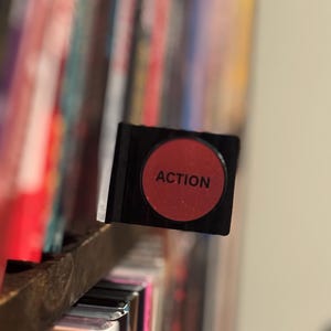May include: A black rectangular divider tab featuring a circular red center with the word ACTION printed in black capital letters. The tab is positioned horizontally, extending from a shelf filled with various colorful media cases.