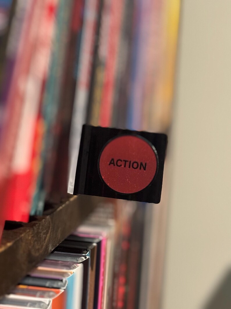 May include: A black rectangular divider tab featuring a circular red center with the word ACTION printed in black capital letters. The tab is positioned horizontally, extending from a shelf filled with various colorful media cases.