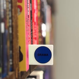 May include: A white rectangular shelf divider featuring a prominent blue circular sticker with the word DRAMA printed in black capital letters, positioned between vertical books on a wooden shelf to categorize the collection by genre.