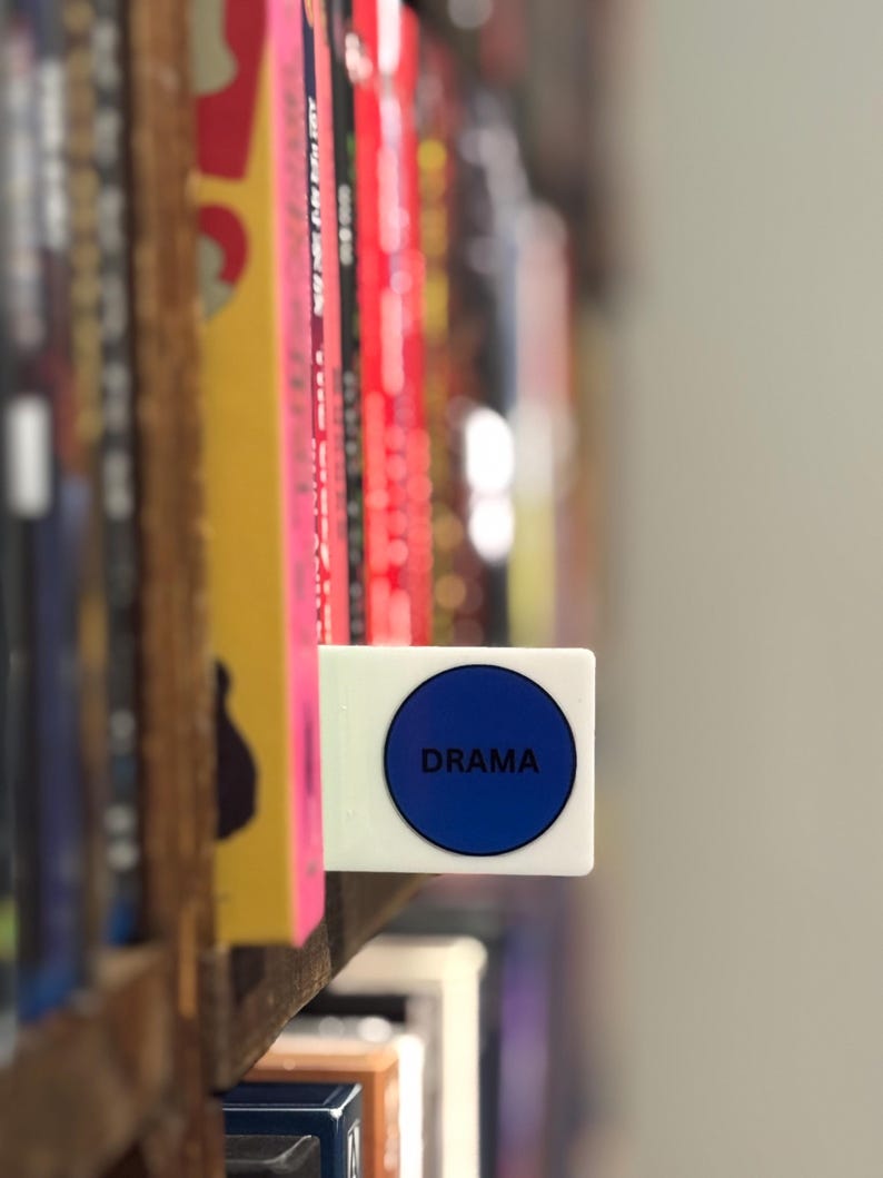 May include: A white rectangular shelf divider featuring a prominent blue circular sticker with the word DRAMA printed in black capital letters, positioned between vertical books on a wooden shelf to categorize the collection by genre.