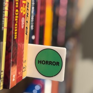 May include: A white rectangular shelf divider featuring a prominent green circular sticker with the word HORROR printed in bold black capital letters. The divider is positioned between two vertical media cases on a shelf, serving as a label.