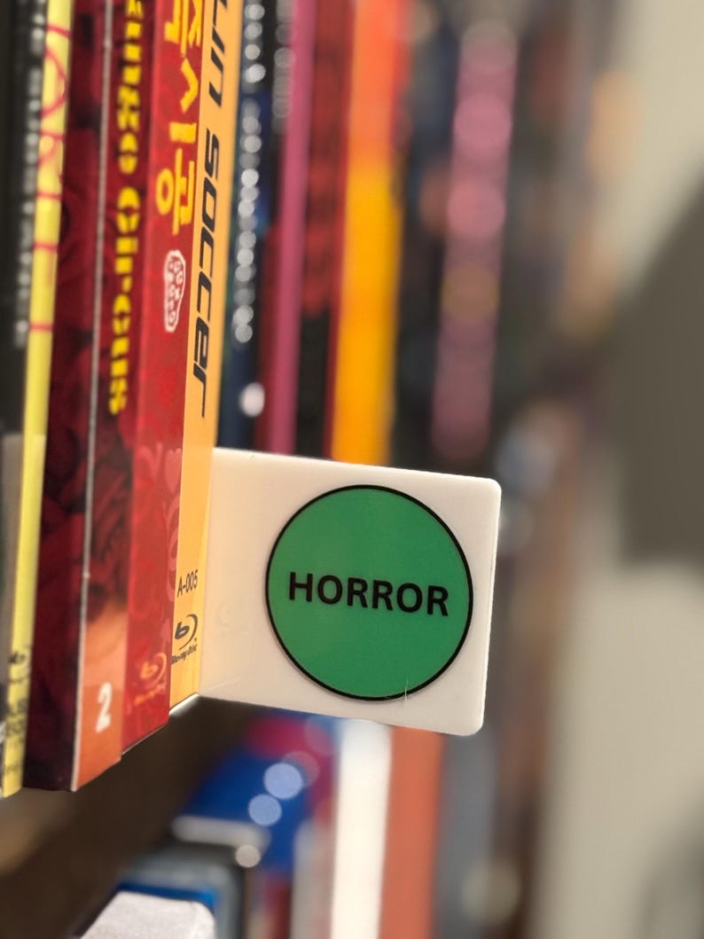 May include: A white rectangular shelf divider featuring a prominent green circular sticker with the word HORROR printed in bold black capital letters. The divider is positioned between two vertical media cases on a shelf, serving as a label.