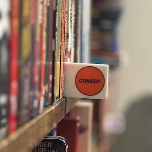 May include: A white rectangular shelf divider featuring a circular orange sticker with the word COMEDY printed in black capital letters. The divider is positioned between rows of DVD cases on a wooden shelf, serving as a genre label.