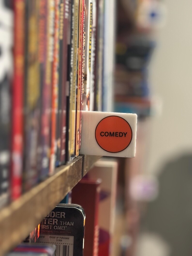 May include: A white rectangular shelf divider featuring a circular orange sticker with the word COMEDY printed in black capital letters. The divider is positioned between rows of DVD cases on a wooden shelf, serving as a genre label.