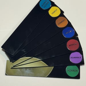May include: A set of seven black rectangular fan-shaped markers, each featuring a circular colored label with black text. The labels are labeled Science Fiction, Family, Comedy, Drama, Action, Romance, and Horror in their respective colors.