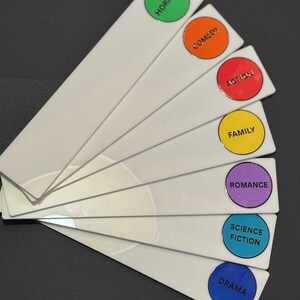 May include: A set of seven white fan-shaped bookmarks arranged in a stack. Each bookmark features a circular colored label at the top with black text reading Horror, Comedy, Action, Family, Romance, Science Fiction, and Drama respectively.