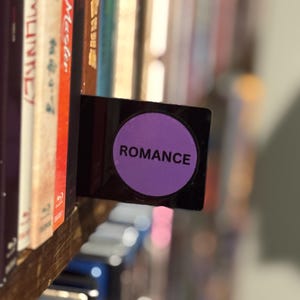 May include: A black rectangular shelf divider featuring a prominent purple circular label in the center. The word ROMANCE is printed in bold black capital letters across the middle of the purple circle, positioned between rows of various media cases.