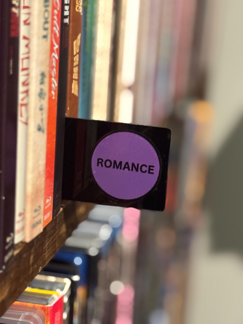 May include: A black rectangular shelf divider featuring a prominent purple circular label in the center. The word ROMANCE is printed in bold black capital letters across the middle of the purple circle, positioned between rows of various media cases.