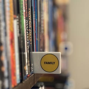 May include: A white rectangular shelf divider featuring a circular yellow sticker with the word FAMILY printed in black capital letters. The divider is positioned between two rows of media cases on a wooden shelf, acting as a label for the collection.