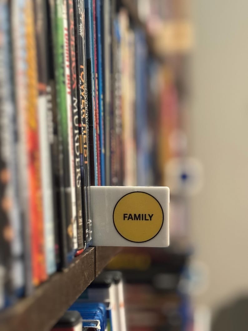 May include: A white rectangular shelf divider featuring a circular yellow sticker with the word FAMILY printed in black capital letters. The divider is positioned between two rows of media cases on a wooden shelf, acting as a label for the collection.