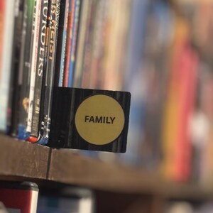 May include: A black rectangular shelf divider featuring a circular yellow sticker with the word FAMILY printed in black capital letters, positioned between rows of various DVD and Blu-ray cases on a wooden shelf in a home media collection.