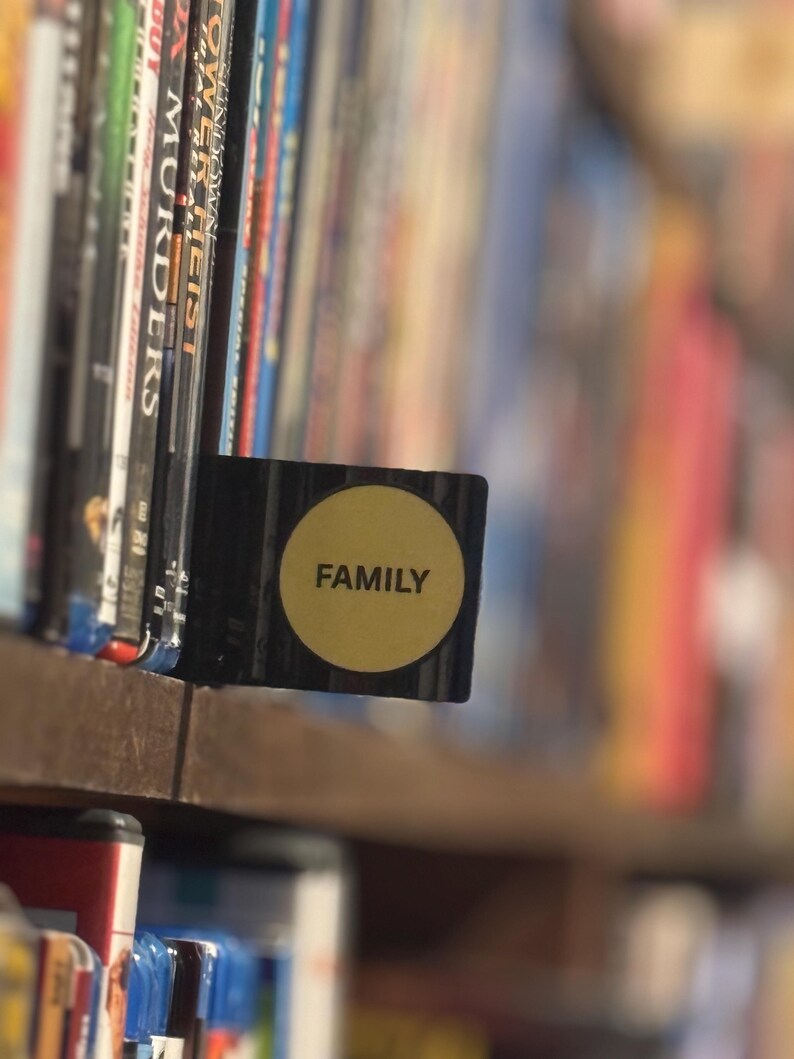 May include: A black rectangular shelf divider featuring a circular yellow sticker with the word FAMILY printed in black capital letters, positioned between rows of various DVD and Blu-ray cases on a wooden shelf in a home media collection.