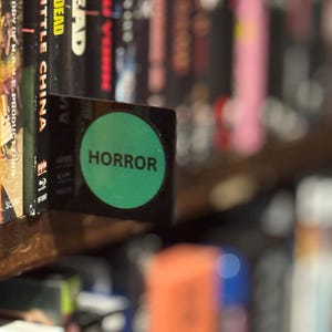 May include: A black rectangular shelf divider featuring a prominent green circular sticker with the word HORROR printed in black capital letters. The divider is positioned between movie cases on a shelf to categorize the collection.