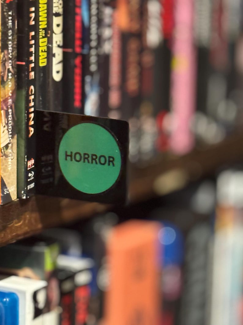 May include: A black rectangular shelf divider featuring a prominent green circular sticker with the word HORROR printed in black capital letters. The divider is positioned between movie cases on a shelf to categorize the collection.