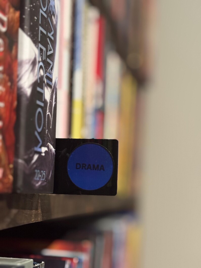 May include: A black rectangular shelf divider featuring a circular blue insert with the word DRAMA printed in black text. The divider is positioned between books on a shelf, serving as a label to categorize the collection of media items.