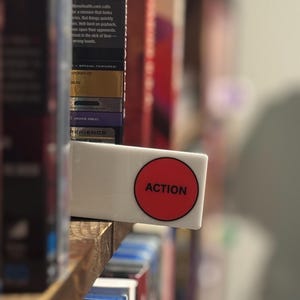 May include: A rectangular white shelf divider featuring a prominent red circle with the word ACTION printed in black capital letters. The divider is positioned between rows of movie cases on a wooden shelf, serving as a clear organizational label.