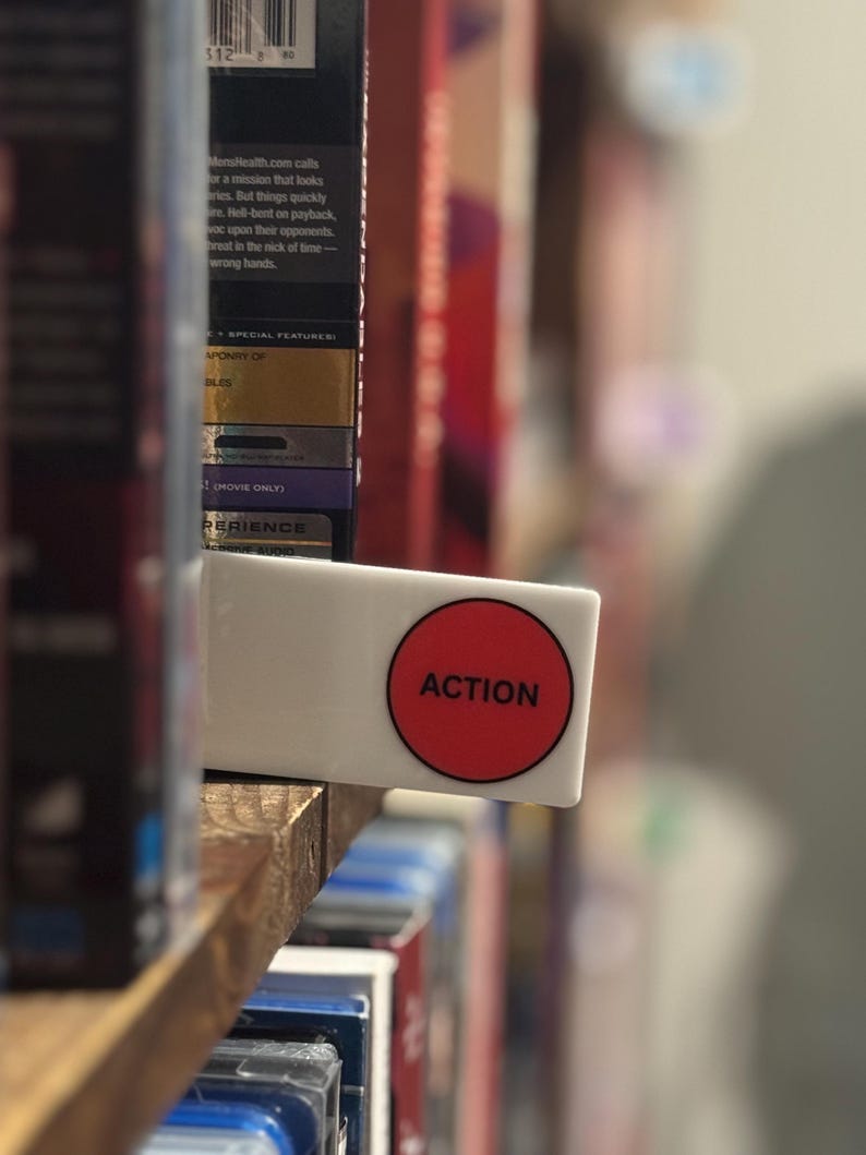 May include: A rectangular white shelf divider featuring a prominent red circle with the word ACTION printed in black capital letters. The divider is positioned between rows of movie cases on a wooden shelf, serving as a clear organizational label.