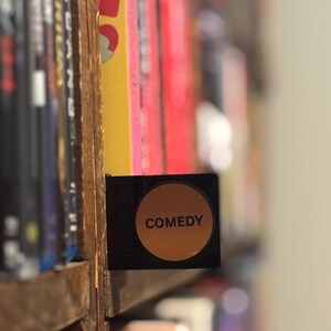 May include: A black rectangular shelf divider featuring a circular orange center with the word COMEDY printed in black capital letters. The divider is positioned between rows of media cases on a wooden shelf, serving as a genre label.