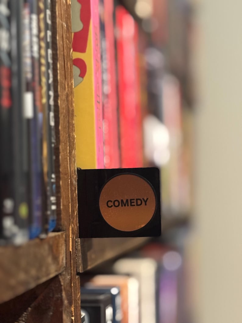 May include: A black rectangular shelf divider featuring a circular orange center with the word COMEDY printed in black capital letters. The divider is positioned between rows of media cases on a wooden shelf, serving as a genre label.