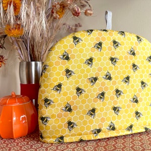 May include: A yellow and black tea cozy with a honeycomb pattern and bee design. The cozy is made of fabric and has a white string loop for hanging.