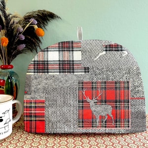 May include: A patchwork tea cozy with a deer silhouette on a red plaid square. The cozy features various gray and red plaid and textured fabrics. A vase with flowers and a mug are in the background.