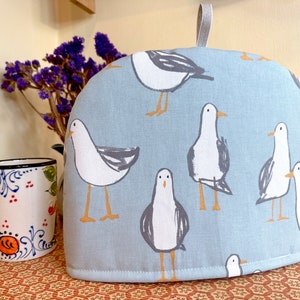 May include: A light blue teapot cozy with a white and gray seagull pattern. The cozy has a loop handle and is made of fabric.