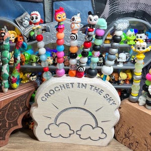 May include: A wooden stand shaped like a cloud with a sun and rainbow design. The stand is engraved with the text "Crochet in the Sky".  The stand holds several colorful beaded pens.