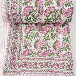 May include: A white quilt with a pink floral pattern and green leaves. The quilt has a pink and white striped border. The floral design features large pink flowers and smaller green leaves. The quilt is folded over.