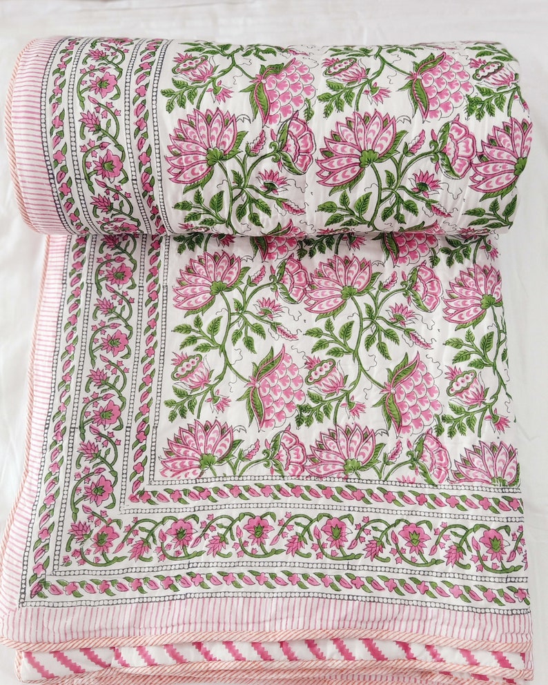 May include: A white quilt with a pink floral pattern and green leaves. The quilt has a pink and white striped border. The floral design features large pink flowers and smaller green leaves. The quilt is folded over.