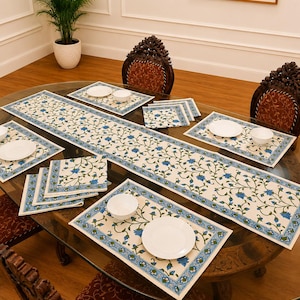 May include: A dining table set featuring a long rectangular table runner, four rectangular placemats, and several folded napkins. Each piece is off-white with a repeating blue floral vine pattern and a blue border with small geometric details.