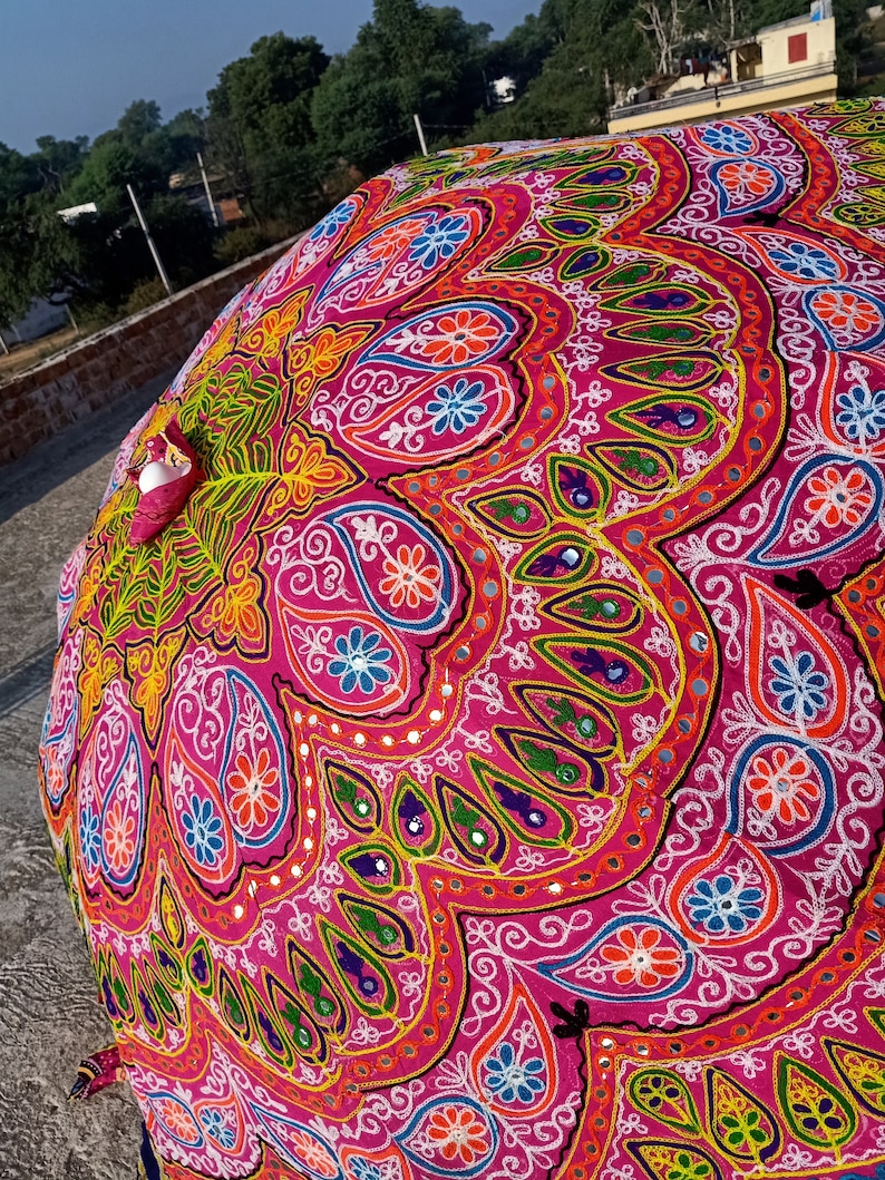 Może przedstawiać: R&oacute;żowy, okrągły parasol z tkaniny z kolorowymi haftami i akcentami lustrzanymi. Parasol jest ozdobiony wzorami paisley i motywami kwiatowymi.