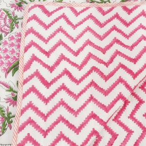 May include: Close-up of a quilted textile with a white background and a vibrant pink zigzag pattern. The edge is trimmed with a thin pink and white striped border. Another section of the textile features a floral design with pink flowers and green leaves.