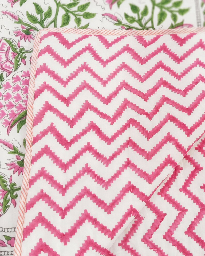 May include: Close-up of a quilted textile with a white background and a vibrant pink zigzag pattern. The edge is trimmed with a thin pink and white striped border. Another section of the textile features a floral design with pink flowers and green leaves.