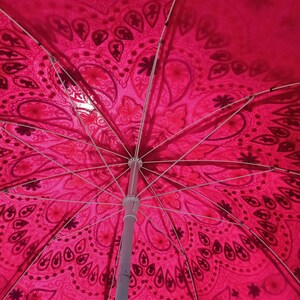 Może przedstawiać: R&oacute;żowy i biały parasol plażowy z wzorem i białą metalową ramą. Parasol jest otwarty, a wnętrze baldachimu jest widoczne.